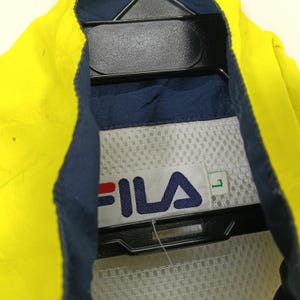 May include: Close-up of a yellow and navy blue Fila jacket. The jacket features the Fila logo in red and navy blue. The jacket is hanging on a black hanger. The size is L.