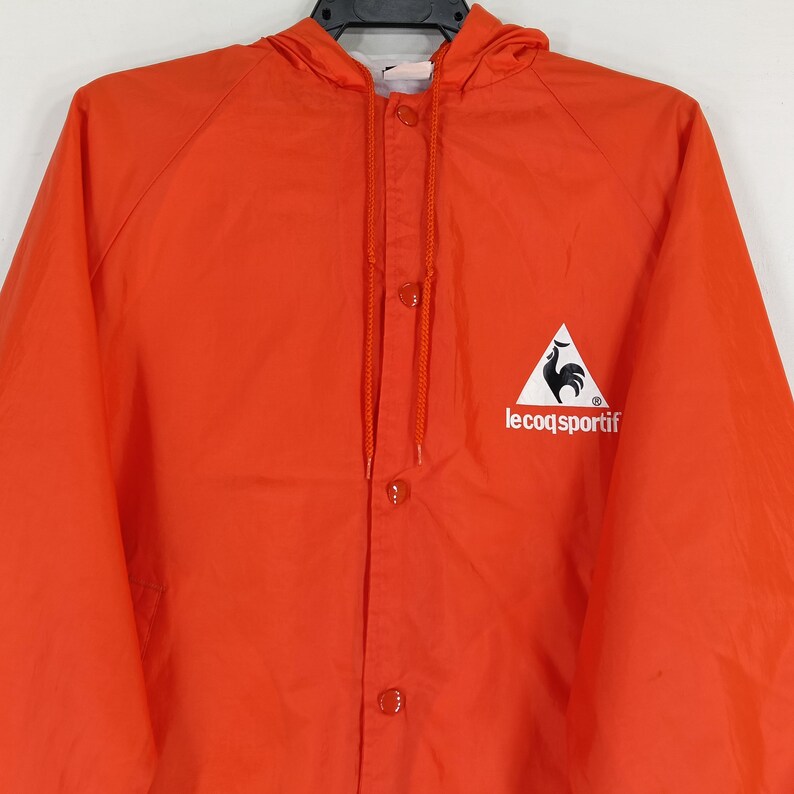 K&ouml;nnte beinhalten: Leuchtend orangefarbene Kapuzenjacke mit Druckknopfverschluss. Die Jacke hat ein wei&szlig;es Dreieckslogo mit einem schwarzen Hahn und dem Text "le coq sportif". Sie hat eine Kapuze mit Kordelzug und ist aus leichtem Material gefertigt.