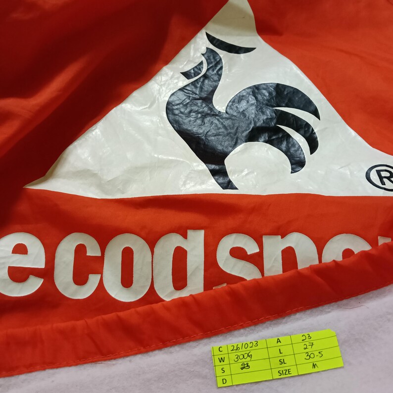K&ouml;nnte beinhalten: Nahaufnahme eines rot-wei&szlig;en Kleidungsst&uuml;cks mit dem Text "ecodsno" in Wei&szlig;. Oben befindet sich ein gro&szlig;es wei&szlig;es Dreieck mit einem schwarzen Hahn-Logo. Ein kleines gelbes Etikett mit Ma&szlig;en befindet sich unten rechts.