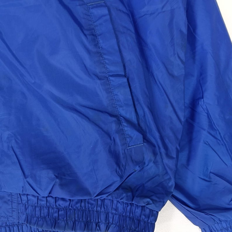 May include: A close-up of a vibrant blue jacket. The jacket features a pocket on the upper left side and an elastic waistband. The fabric has a slight sheen, suggesting a nylon or similar material. The jacket is likely part of a sportswear or casual wear collection.