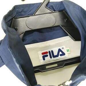 May include: Navy blue FILA jacket with a white interior and a black hanger. The FILA logo is displayed on a white tag with red and blue accents. The jacket is size M.