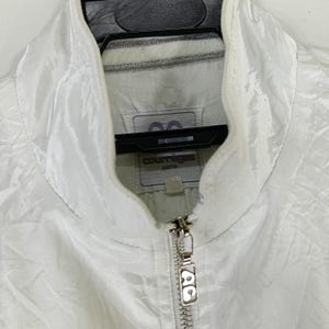 May include: White Courr&egrave;ges Paris jacket with a high collar and a full zipper. The jacket has a subtle sheen and a small logo tag near the collar. The zipper pull features the Courr&egrave;ges logo.