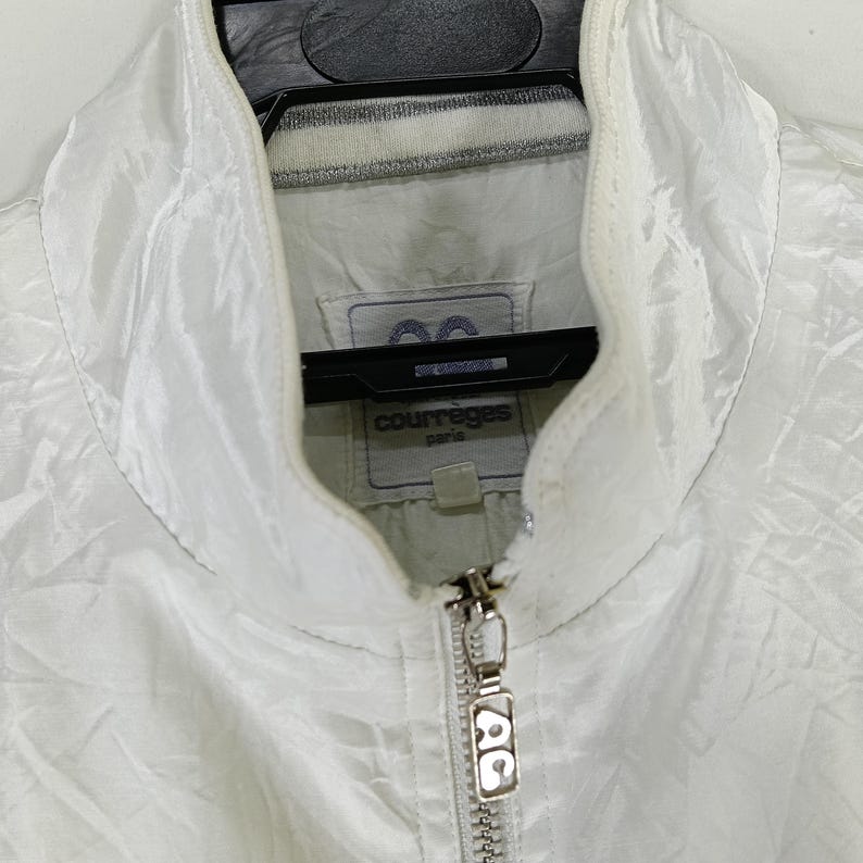 May include: White Courr&egrave;ges Paris jacket with a high collar and a full zipper. The jacket has a subtle sheen and a small logo tag near the collar. The zipper pull features the Courr&egrave;ges logo.
