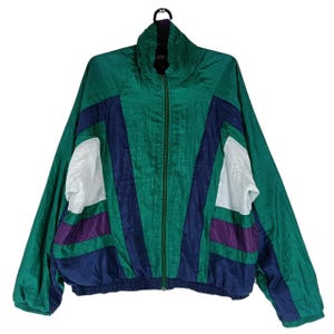 Könnte beinhalten: Eine Vintage-Windjacke in Grün, Blau, Weiß und Lila mit durchgehendem Reißverschluss. Die Jacke hat einen hohen Kragen und ein Colorblock-Design. Sie hängt an einem schwarzen Kleiderbügel.
