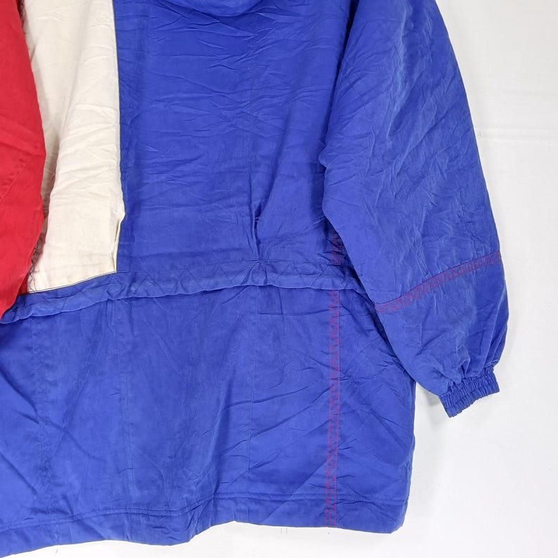 May include: A vintage blue, red, and white anorak jacket. The jacket features a color block design with a red panel on the left, a white panel in the center, and a blue panel on the right. The jacket has a hood and a drawstring at the waist.