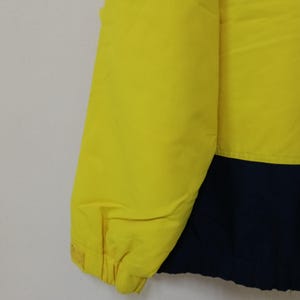 Puede incluir: Primer plano de una chaqueta amarilla y azul marino. La chaqueta est&aacute; hecha de un material liso, posiblemente nailon. La parte amarilla est&aacute; en la parte superior izquierda y el azul marino en la inferior derecha. La chaqueta parece ligeramente acolchada.