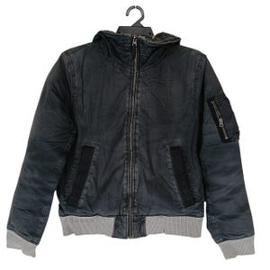 Pode incluir: Um blusão bomber com capuz em cinzento-carvão, com fecho de correr frontal a todo o comprimento, bainha e punhos canelados em cinzento, dois bolsos laterais verticais com rebordo preto e um pequeno bolso utilitário com fecho na manga esquerda.