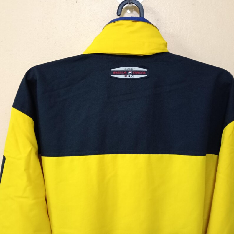 Puede incluir: Chaqueta amarilla y azul marino con cuello azul y un peque&ntilde;o logotipo en la espalda. La chaqueta est&aacute; colgada de una percha negra. La chaqueta tiene un dise&ntilde;o de bloques de color.