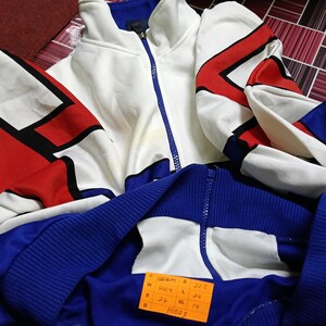 May include: White and blue tracksuit jacket with red and black geometric designs. The jacket has a zipper closure and a size tag that reads "W 4006 A 22.5 S 24 L 24 BL 19 11023".
