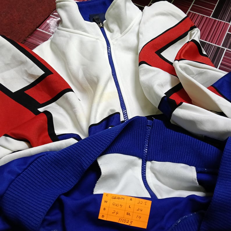 May include: White and blue tracksuit jacket with red and black geometric designs. The jacket has a zipper closure and a size tag that reads "W 4006 A 22.5 S 24 L 24 BL 19 11023".