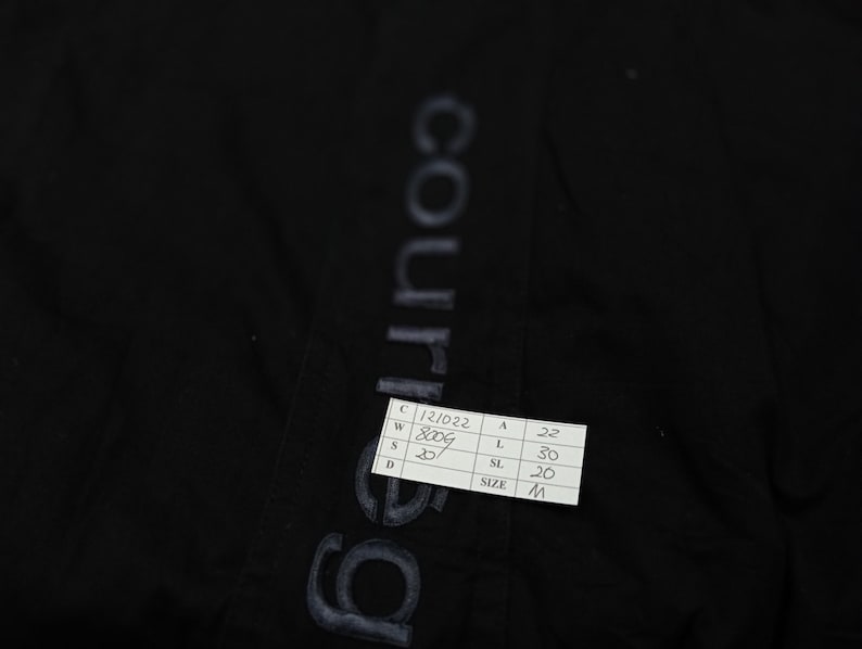 May include: Black garment with the word "courreges" printed vertically in a faded, light color. A small white tag with black text is attached, displaying measurements and the size "M".