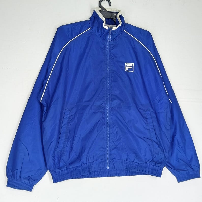 May include: A blue zip-up jacket with white trim and a white collar. The jacket features the brand logo on the chest. The jacket has two front pockets and a full-length zipper. The jacket is made of a lightweight material.
