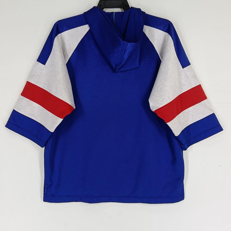 K&ouml;nnte beinhalten: Ein blauer Kapuzenpullover mit wei&szlig;en und roten Streifen an den &Auml;rmeln. Der Pullover hat eine lockere Passform und einen l&auml;ssigen Stil.