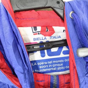 May include: Close-up of a blue and red jacket with a white label. The label reads "BIELLA ITALIA" and features the brand's logo. The jacket has a zipper and a drawstring hood. The text "ILA opera nel mondo dell sport in cinque e foundamental: abito da" is also visible.