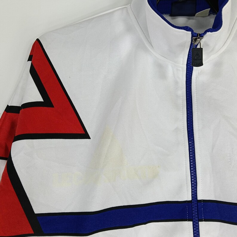 May include: White track jacket with red, black, and blue geometric patterns. The jacket has a zipper closure and a stand-up collar.