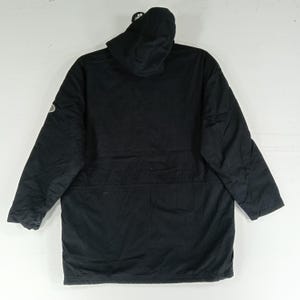 May include: A black hooded parka jacket with long sleeves. The jacket has a drawstring waist and a small logo on the left sleeve. The hood is attached and the jacket is made of a durable material.