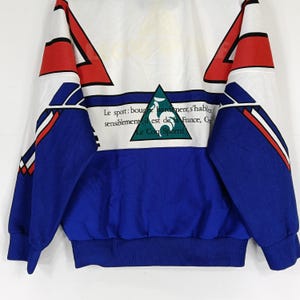 May include: A blue and white track jacket with red and white accents. The jacket has a large green rooster logo on the front with the text "Le sport: bougez hardiment, s'habillez sensiblement, il est de la France, Cr Le Coq Sportif".