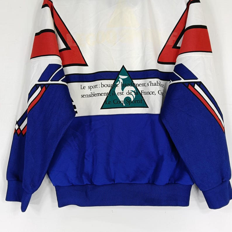 May include: A blue and white track jacket with red and white accents. The jacket has a large green rooster logo on the front with the text "Le sport: bougez hardiment, s'habillez sensiblement, il est de la France, Cr Le Coq Sportif".