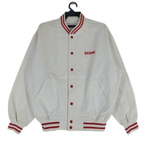 May include: Cream-colored bomber jacket with red accents. Features a red and white striped collar, cuffs, and hem. The jacket has red buttons and the word "SCENE" embroidered in red on the chest. Includes two front pockets.