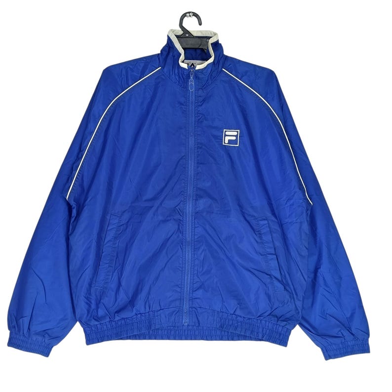 May include: A royal blue zip-up jacket with white trim and a white logo on the chest. The jacket has a high collar and a full-length zipper. The jacket is made of a lightweight material, perfect for layering.