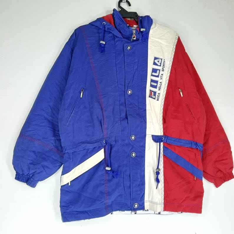May include: A vintage Fila jacket in blue, red, and white. The jacket features a hooded design, a zip-up front, and a drawstring waist. The Fila logo is prominently displayed on the front. The jacket has a sporty, retro aesthetic.