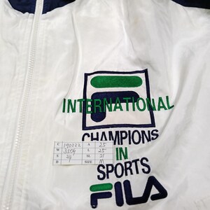 May include: White and navy blue FILA jacket with a full zipper. The jacket features the text "INTERNATIONAL CHAMPIONS IN SPORTS" and the FILA logo in green and navy blue.