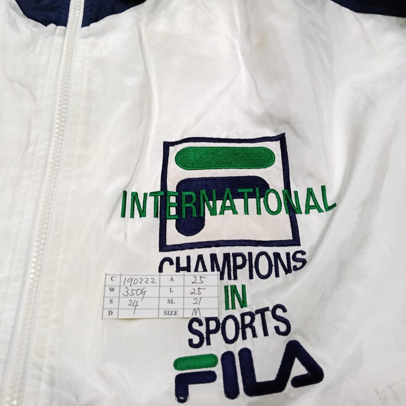 May include: White and navy blue FILA jacket with a full zipper. The jacket features the text "INTERNATIONAL CHAMPIONS IN SPORTS" and the FILA logo in green and navy blue.