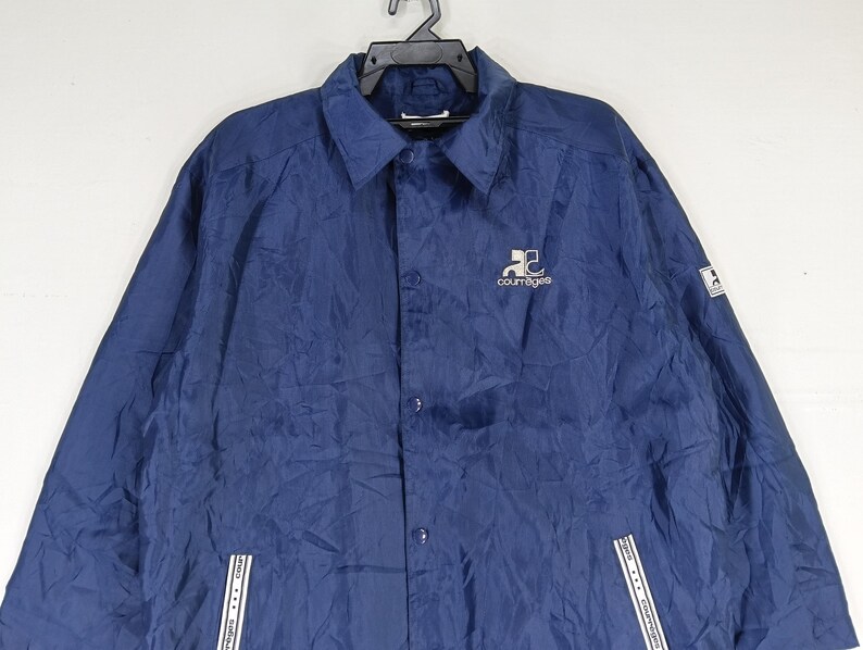 May include: A navy blue jacket with a button-up front and a white logo that says "Couragea" on the left chest. The jacket has two white zippered pockets on the bottom.