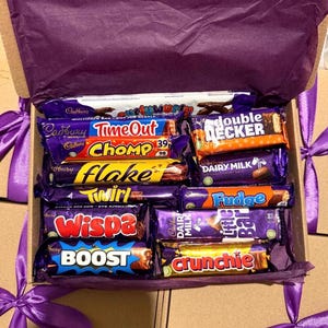 May include: A gift box filled with various Cadbury chocolate bars, including Time Out, Chomp, Flake, Twirl, Wispa, Boost, and Crunchie. The box is lined with purple tissue paper and tied with a purple ribbon.