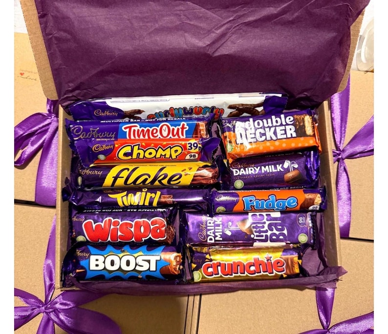 May include: A gift box filled with various Cadbury chocolate bars, including Time Out, Chomp, Flake, Twirl, Wispa, Boost, and Crunchie. The box is lined with purple tissue paper and tied with a purple ribbon.