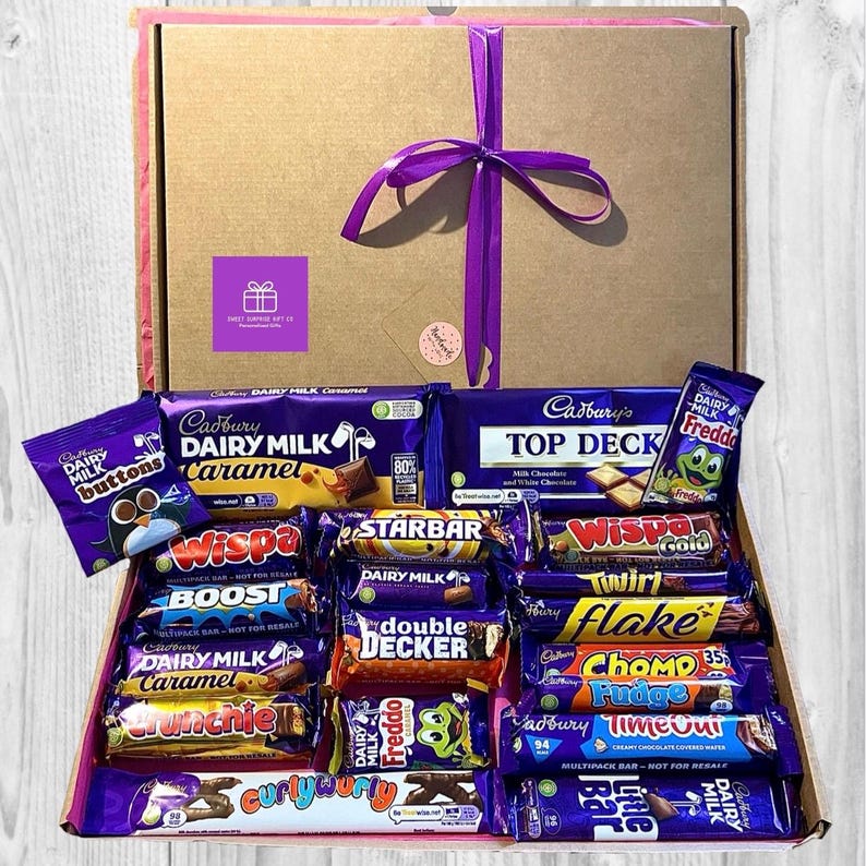 May include: A gift box filled with various Cadbury chocolate bars and candies. The box is brown with a purple ribbon and bow. The assortment includes Dairy Milk, Wispa, Starbar, and Freddo bars, among others. The box has a pink interior.