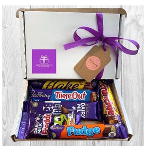 May include: A white gift box tied with a purple ribbon, filled with various Cadbury chocolate bars. The box has a purple sticker with a gift icon and the text "Sweet Surprise Gift Co". A brown tag with the word "Handmade" is attached.