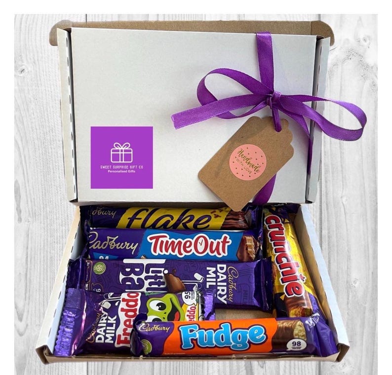 May include: A white gift box tied with a purple ribbon, filled with various Cadbury chocolate bars. The box has a purple sticker with a gift icon and the text "Sweet Surprise Gift Co". A brown tag with the word "Handmade" is attached.