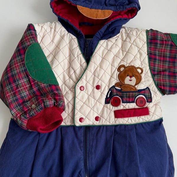 May include: A navy blue and cream-coloured baby snowsuit with a quilted front panel and plaid sleeves. The snowsuit features a hood, a teddy bear applique, and red accents. The suit has a full-length zip.