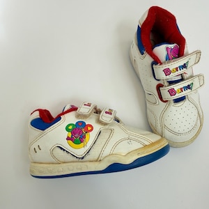 Könnte beinhalten: Ein Paar weiße Turnschuhe mit Barney-dem-Dinosaurier-Grafiken. Die Schuhe haben blaue Sohlen, ein rotes Innenfutter und zwei Klettverschlüsse an jedem Schuh, auf denen das Wort Barney in bunten Buchstaben aufgedruckt ist. Die Seiten zeigen ein Barney-Motiv.