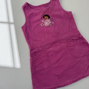 May include: A magenta corduroy dress with an embroidered cartoon character and floral design. The dress has a sleeveless design, two front pockets, and a fitted waist. This children's dress is made of corduroy.