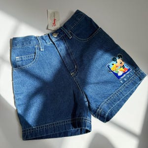 Puede incluir: Shorts vaqueros azules con un gráfico de Mickey Mouse en la pierna derecha. Los shorts tienen un diseño clásico de cinco bolsillos y cierre de botón. La etiqueta de Disney Store está adjunta.