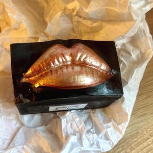 May include: A rectangular black soap bar with a metallic gold lip design. The soap is resting on white tissue paper. The lip design has a glossy finish and is centered on the bar.