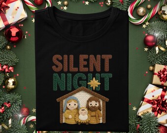 May include: Black t-shirt with the words "SILENT NIGHT" in brown and green knit lettering, and a gold star. Below the text is a knit illustration of the nativity scene. The shirt is surrounded by Christmas decorations.