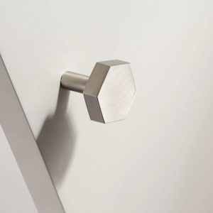Op de afbeelding: Een geborsteld nikkelen kastknop met een geometrisch, zeshoekig ontwerp. De knop steekt uit een wit oppervlak en werpt een schaduw. De hardware heeft een moderne, minimalistische uitstraling.