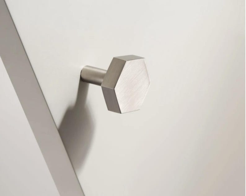 Op de afbeelding: Een geborsteld nikkelen kastknop met een geometrisch, zeshoekig ontwerp. De knop steekt uit een wit oppervlak en werpt een schaduw. De hardware heeft een moderne, minimalistische uitstraling.