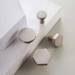 May include: A collection of brushed nickel cabinet hardware, including round and hexagonal knobs. The knobs are in various sizes and shapes, with a modern, minimalist design. The hardware is displayed on a white surface.