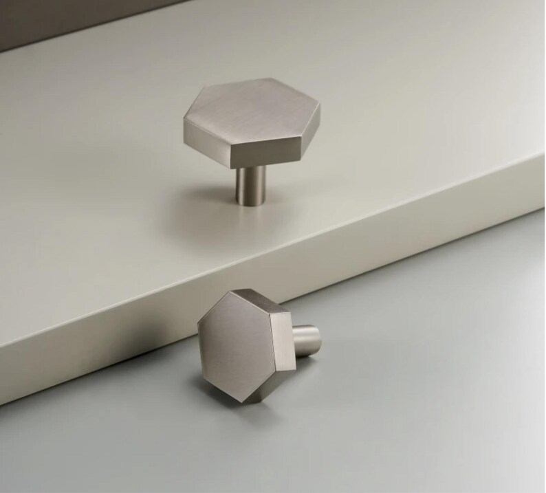 Op de afbeelding: Twee kastknoppen van geborsteld nikkel met een zeshoekig ontwerp. E&eacute;n knop staat rechtop, bevestigd aan een oppervlak, terwijl de andere op een grijs oppervlak ligt. De knoppen hebben een moderne, minimalistische uitstraling.