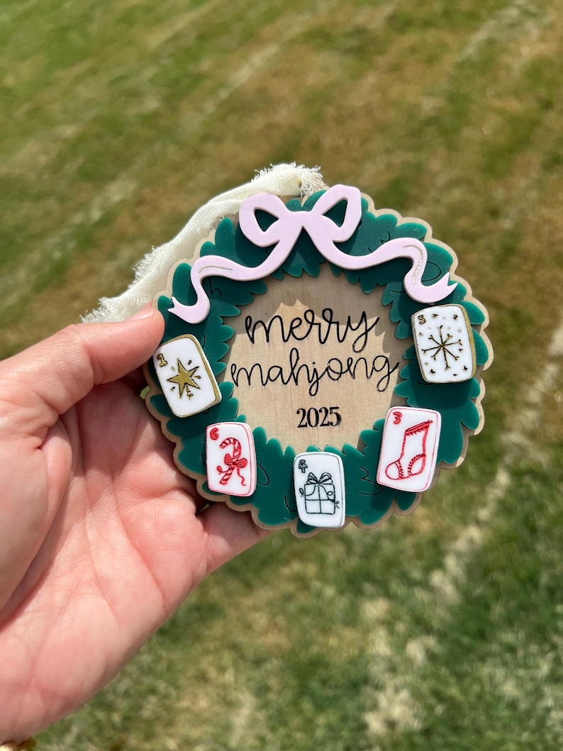 May include: A Christmas ornament with a green wreath design, a pink bow, and the words "merry mahjong 2025". The ornament features six small, colorful card-like images with festive symbols.