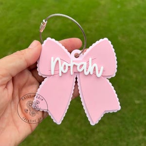 May include: A pink bow-shaped luggage tag with the name "Norah" in white script. The tag has a scalloped edge and a silver-colored metal loop for attachment. The background is green.