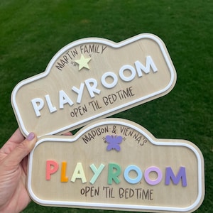 Puede incluir: Dos letreros de madera clara con bordes blancos en relieve. El letrero superior dice MARTIN FAMILY PLAYROOM OPEN 'TIL BEDTIME con una estrella amarilla. El letrero inferior dice MADISON & VIENNA'S PLAYROOM OPEN 'TIL BEDTIME con una mariposa morada.