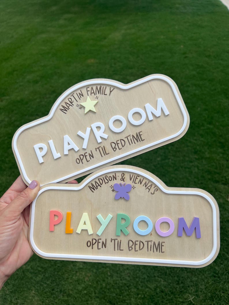 Puede incluir: Dos letreros de madera clara con bordes blancos en relieve. El letrero superior dice MARTIN FAMILY PLAYROOM OPEN 'TIL BEDTIME con una estrella amarilla. El letrero inferior dice MADISON & VIENNA'S PLAYROOM OPEN 'TIL BEDTIME con una mariposa morada.