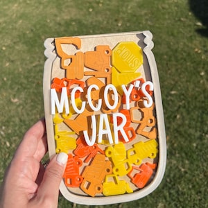 Puede incluir: Letrero de madera en forma de tarro con el texto "MCCOY'S JAR" en blanco. El tarro está lleno de vehículos de construcción de juguete naranjas y amarillos y una señal de stop. El letrero se sostiene sobre un fondo de hierba verde.
