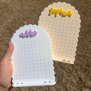May include: Two white earring display stands with scalloped edges. The stand on the left has the name "Addie" in purple lettering. The stand on the right has the name "Sophia" in yellow lettering.