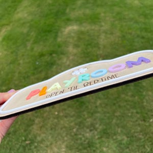 Puede incluir: Un letrero de madera con una superficie clara y un borde oscuro, que presenta la palabra PLAYROOM en letras pastel multicolores en relieve y el texto OPEN TIL BED TIME impreso en negro debajo, con una peque&ntilde;a forma de flor blanca incluida.